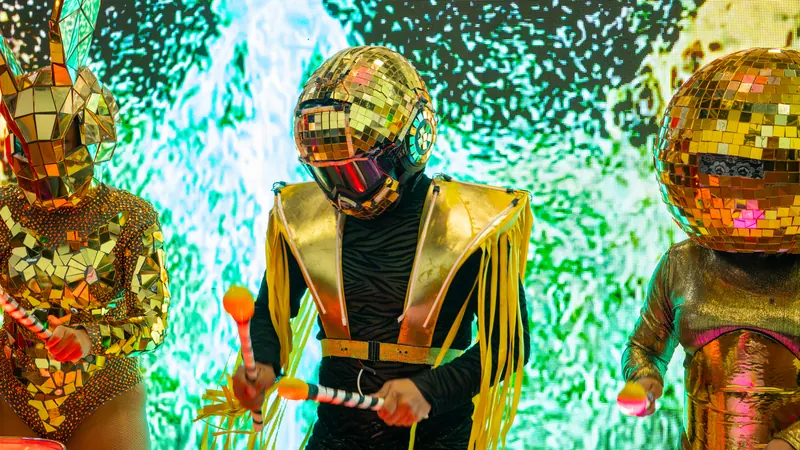 Percusionistas con trajes de espejos dorados tocando con baquetas frente a una pantalla LED
