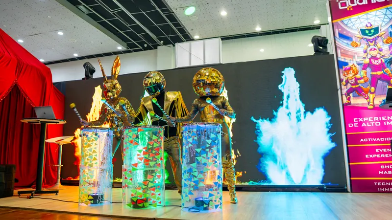 Personajes dorados tocando tambores iluminados frente a una pantalla LED